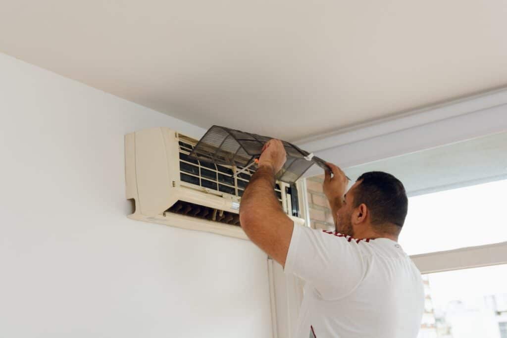 Adult technician man standing on a folding aluminum ladder is taking out dirty filters from air conditioner in apartment, doing service and maintenance, copy space