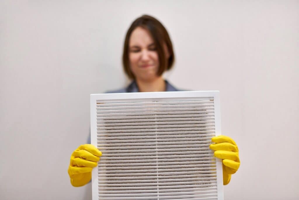 Woman holds ventilation grill with dust filter to clean it. Dirty and dusty white plastic, harmful for health. Cleaning lady with disguised expression, blurred, in protective rubber gloves and uniform